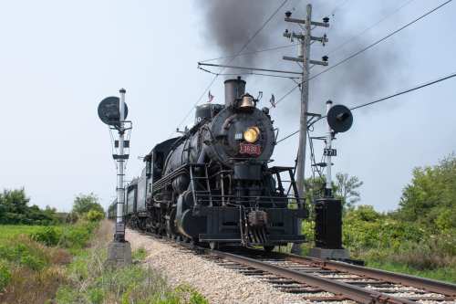 A vintage steam locomotive chugs along a track, emitting smoke, with signal lights and greenery in the background.