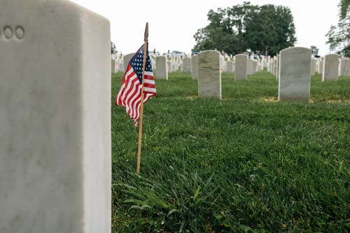 A small American flag stands beside a gravestone in a cemetery, surrounded by rows of white headstones.