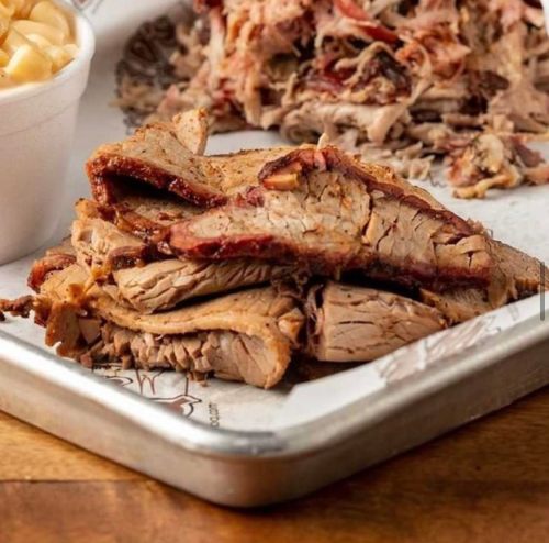 Sliced smoked brisket on a metal tray, with pulled pork and a side of macaroni and cheese in the background.