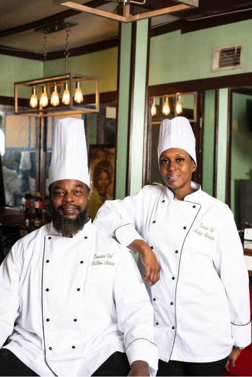 Two chefs in white uniforms and tall hats pose together in a restaurant setting, smiling confidently.