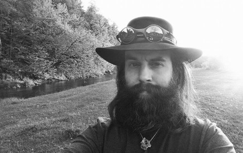 A bearded man in a hat and goggles stands by a river, surrounded by trees, in a black and white photo.