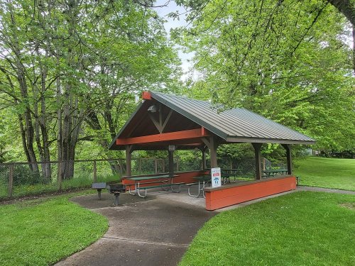 A covered picnic shelter with benches, surrounded by green trees and grass in a park setting.