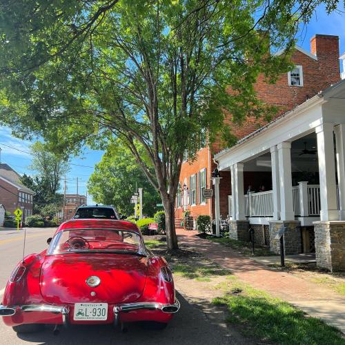 A red vintage car parked on a tree-lined street near a brick house with a porch.