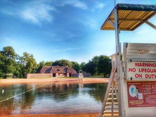 A lifeguard stand by a calm lake with a sandy beach and a building in the background under a clear blue sky.