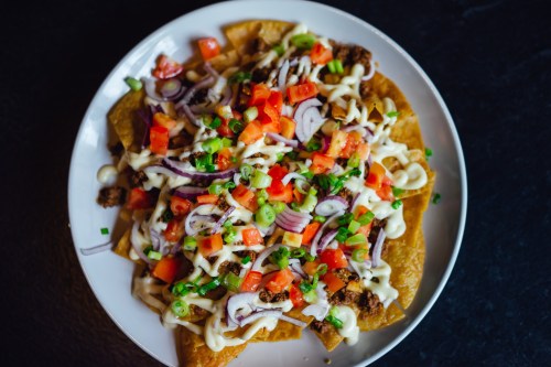 A plate of nachos topped with cheese, diced tomatoes, onions, green onions, and a drizzle of sauce.