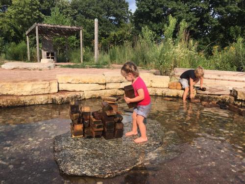 Two children play in a shallow stream, building with wooden blocks on a rock surrounded by greenery.