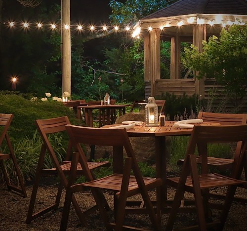 A cozy outdoor dining area with wooden tables, chairs, and string lights, surrounded by greenery at night.