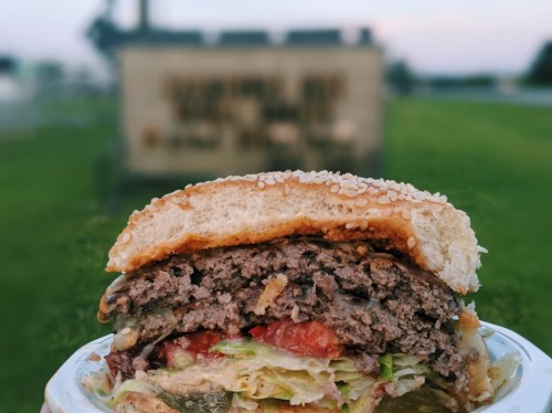 Close-up of a juicy burger with lettuce, tomatoes, and pickles, set against a blurred outdoor sign in the background.