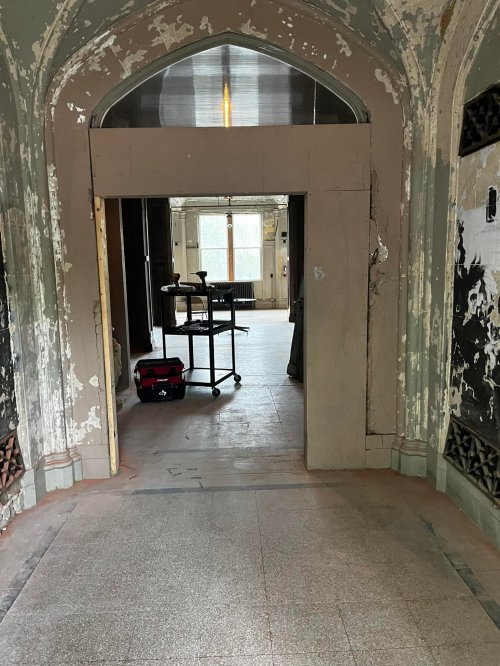A dimly lit hallway with peeling paint, leading to a room with a cart and visible windows in the background.