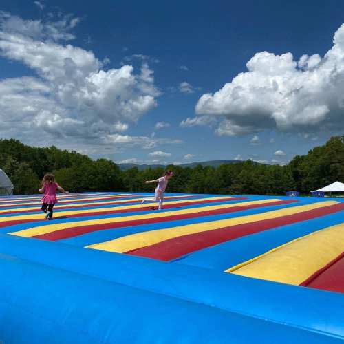 Two children play on a large inflatable bounce pad under a blue sky with fluffy clouds and green trees in the background.