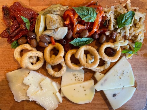 A wooden board featuring a variety of cheeses, olives, sun-dried tomatoes, artichokes, roasted peppers, and breadsticks.