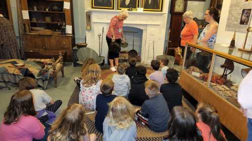 A group of children sits on the floor, watching a woman demonstrate an activity in a museum setting.