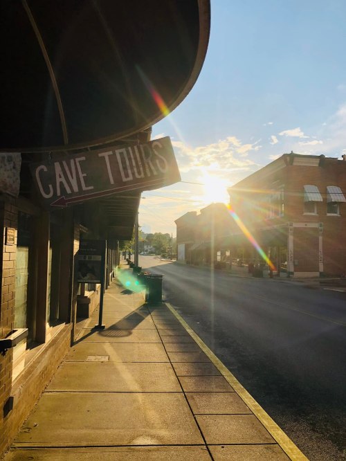 A street scene at sunset with a sign for "Cave Tours" and buildings lining the road. Sunlight creates a lens flare effect.