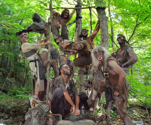 A group of people in primitive costumes, covered in mud, posing playfully among trees in a forest setting.