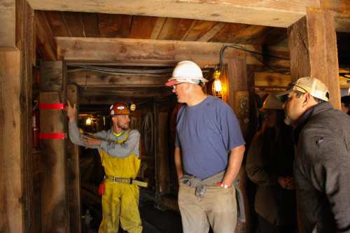 Two men observe a miner in a hard hat demonstrating equipment inside a wooden mine tunnel.