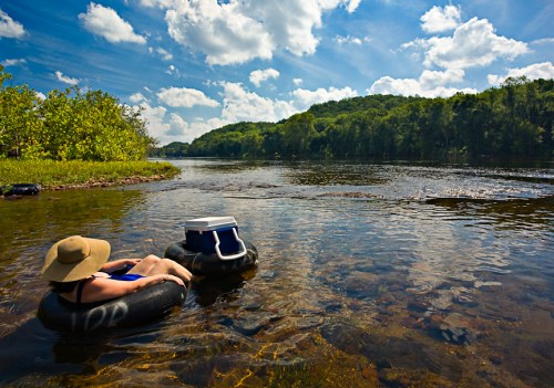 A person relaxes on an inner tube in a river, surrounded by trees and a bright blue sky with fluffy clouds.