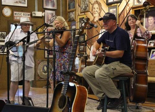 A group of musicians performs on stage, featuring a guitarist, a fiddler, and a bassist, surrounded by framed photos.