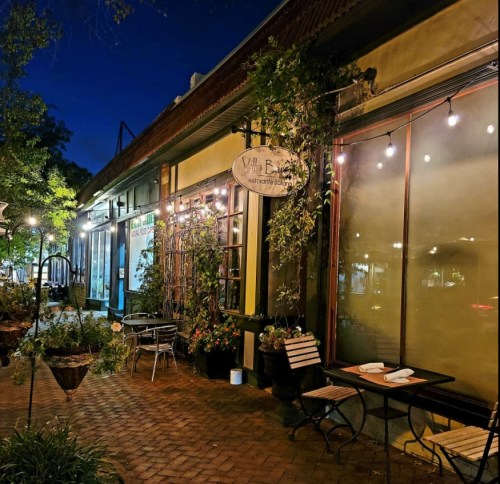 A cozy outdoor dining area at night, featuring string lights, potted plants, and empty tables outside a restaurant.
