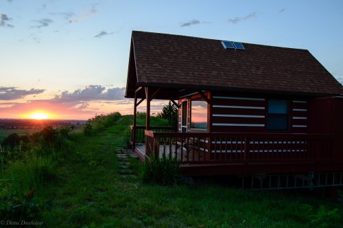 A cozy cabin with a porch overlooks a vibrant sunset, surrounded by lush greenery and open fields.