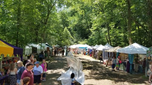 A vibrant outdoor market with colorful tents, people browsing, and lush greenery surrounding the pathway.