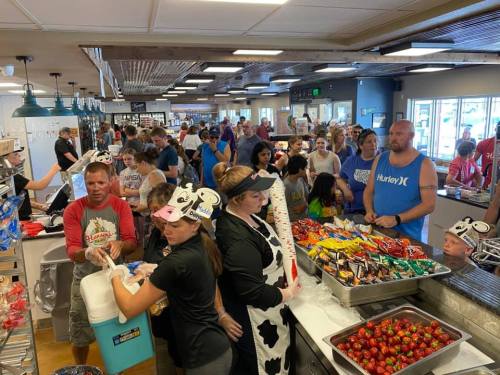 A busy cafeteria scene with a long line of people being served food, including staff in cow-themed attire.