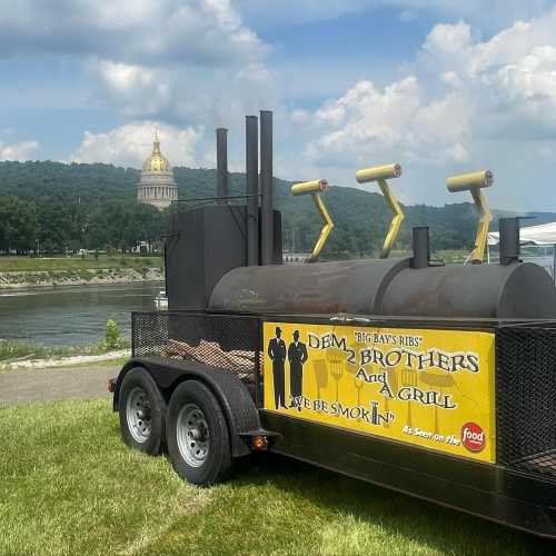 A food trailer with a sign reading "Big Ray's Ribs" near a river, with a state capitol building in the background.