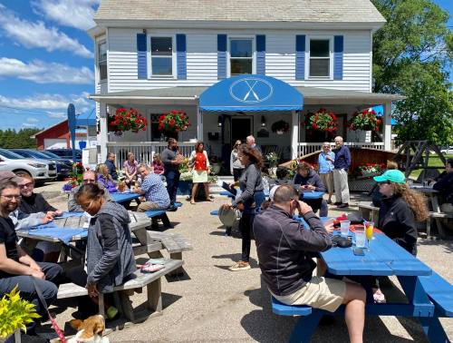 A busy outdoor café with people dining at picnic tables, surrounded by flowers and a blue sky.