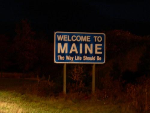 A nighttime photo of a blue welcome sign for Maine, stating, "The Way Life Should Be."