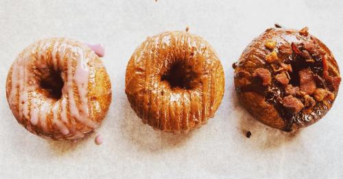 Three donuts on parchment paper: one with pink glaze, one glazed, and one topped with chocolate and bacon bits.