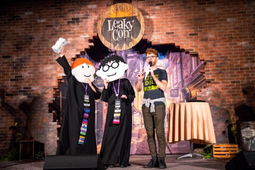Two people in large puppet costumes stand on stage at LeakyCon, while a speaker holds a microphone nearby.