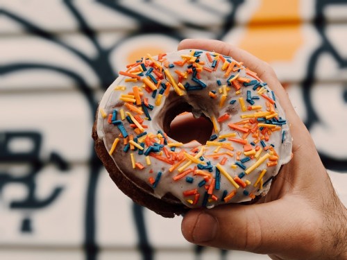 A hand holding a donut topped with white icing and colorful sprinkles against a graffiti background.
