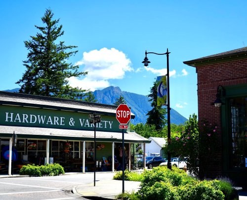 A quaint hardware store with a stop sign, surrounded by greenery and mountains under a clear blue sky.
