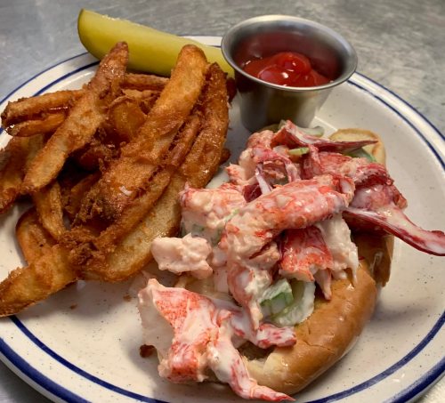 A plate with a lobster roll, crispy fries, a pickle, and a small cup of ketchup.
