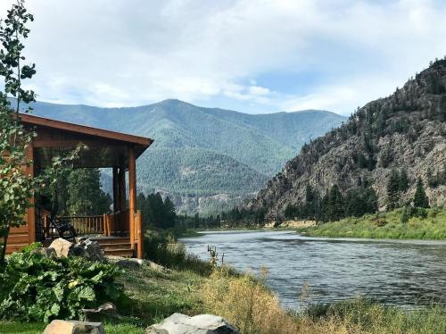 A serene riverside view with a wooden cabin, mountains in the background, and lush greenery along the water's edge.