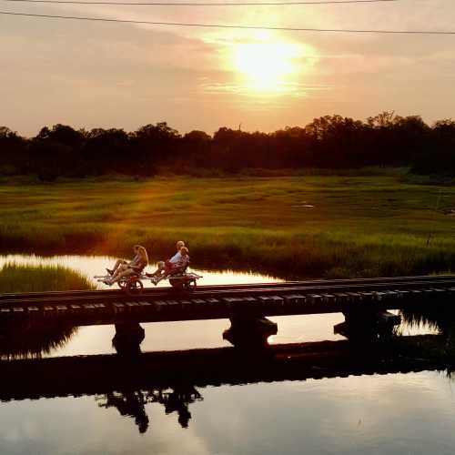 Two people sit on a cart crossing a wooden bridge at sunset, surrounded by lush greenery and calm waters.