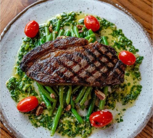 Grilled steak on a bed of green beans and pesto, garnished with cherry tomatoes and pecans on a textured plate.