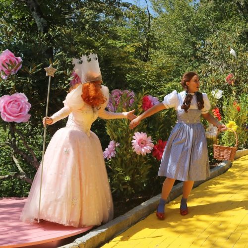 A whimsical scene with a character in a pink gown and crown holding hands with a girl in a blue dress on a yellow path.