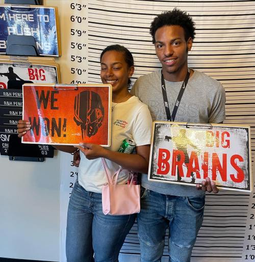 Two young people smile while holding signs that say "We Won!" and "Big Brains!" in front of a height chart backdrop.