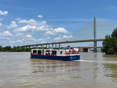 A boat with passengers navigates a river under a large suspension bridge on a sunny day with scattered clouds.
