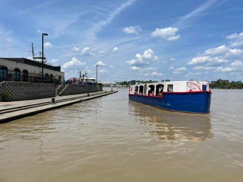 A blue and white boat approaches a dock along a river, with a cloudy sky and buildings in the background.