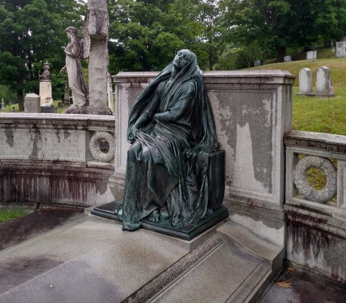 A weathered bronze statue of a seated figure draped in cloth, surrounded by a cemetery landscape.