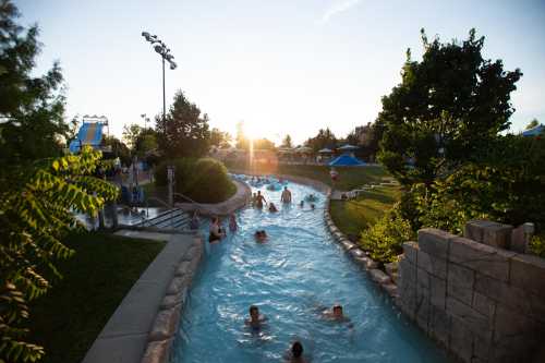 A lazy river at a water park with people floating, surrounded by greenery and a sunset in the background.