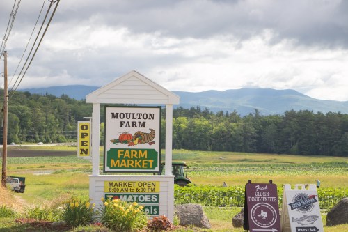 Sign for Moulton Farm Market with mountains in the background, featuring hours and fresh produce offerings.