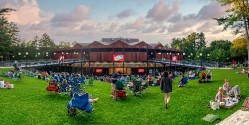 A crowd gathers on a grassy area with chairs, enjoying an outdoor event at a venue surrounded by trees.