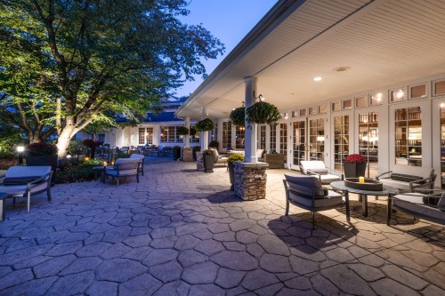 A cozy outdoor patio with stone flooring, seating, and lush greenery, illuminated by soft evening lights.