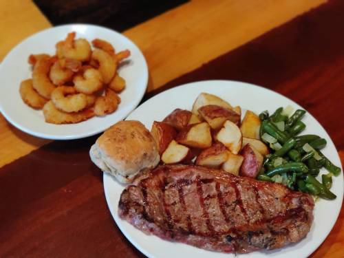 A plate with grilled steak, roasted potatoes, green beans, and a biscuit, alongside a bowl of fried shrimp.
