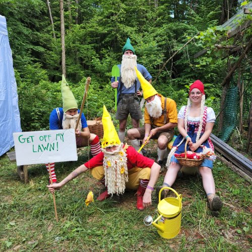 A group of five people dressed as garden gnomes, holding a sign that says "GET OFF MY LAWN!" in a forested area.