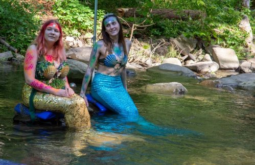 Two mermaids with colorful body paint sit on rocks in a stream, surrounded by greenery.