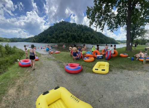 People enjoying a sunny day by a river, surrounded by inflatable tubes and a lush green hillside.