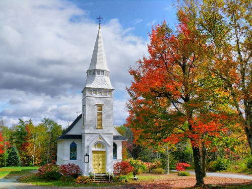 A quaint white church with a tall steeple, surrounded by vibrant autumn foliage and a cloudy sky.